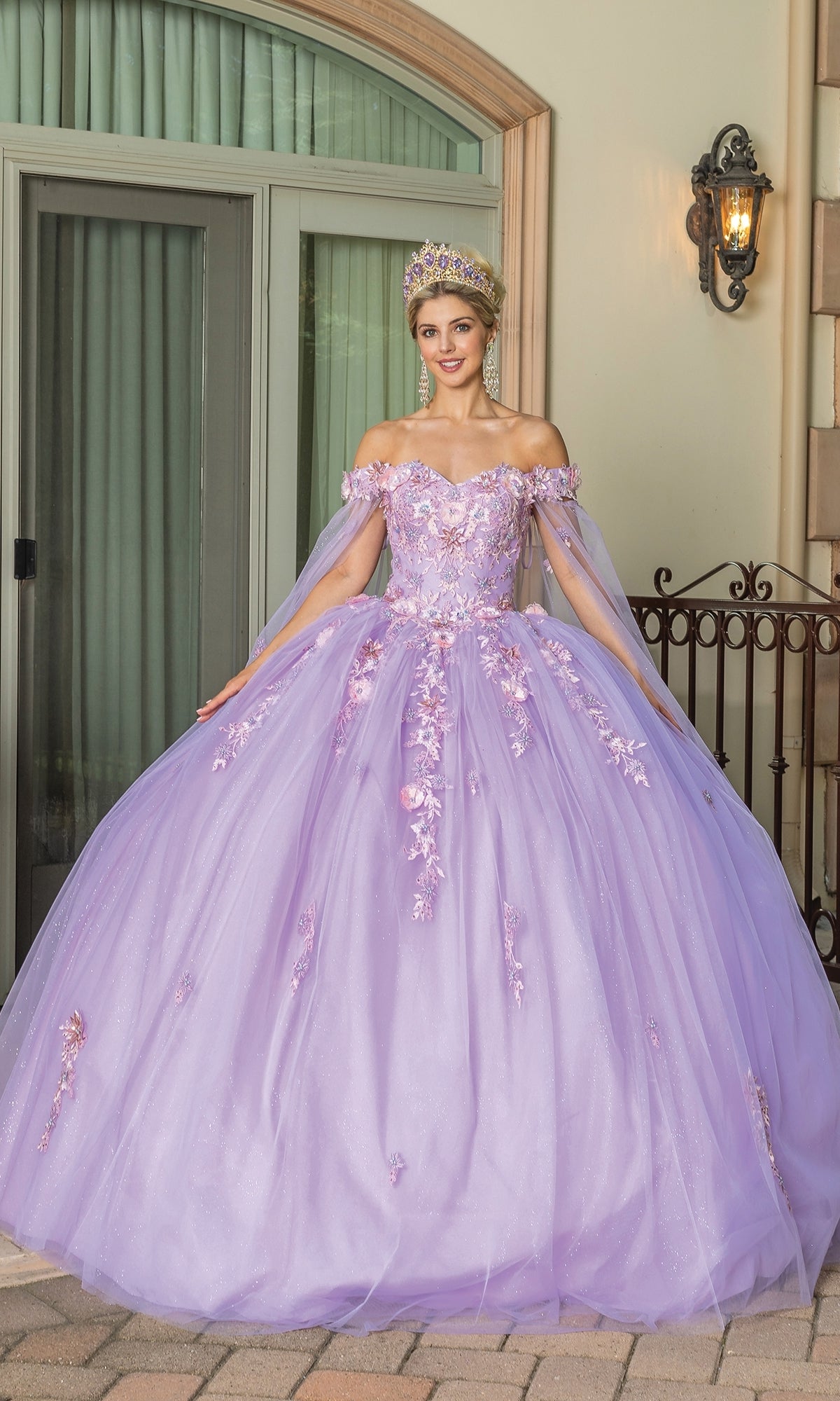 A woman in the Quinceanera Dress 1714 By Dancing Queen, a pastel purple ball gown with floral details and a tiara, stands before a building with large windows and a wrought iron fence.